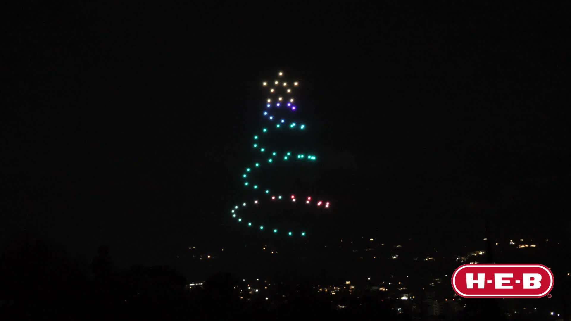 Enjambre de drones de luz formando una figura navideña y el logo de H·E·B en el cielo nocturno
